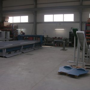 Wnętrze hali produkcyjnej z maszynami CNC, regałami narzędziowymi, pracownikami przy maszynach i gotowymi elementami na podło