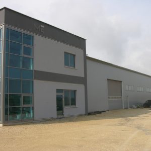 Nowoczesny budynek przemysłowy z szarą fasadą, dużymi okna­mi narożnymi, drzwiami wjazdowymi i parkingiem. Piaszczyste podłoż