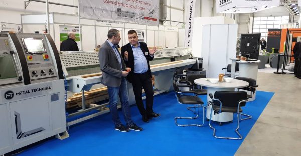 Dwaj mężczyźni w biurowych ubraniach rozmawiają na niebieskiej podłodze targu przemysłowego. W tle maszyny CNC i stanowiska w