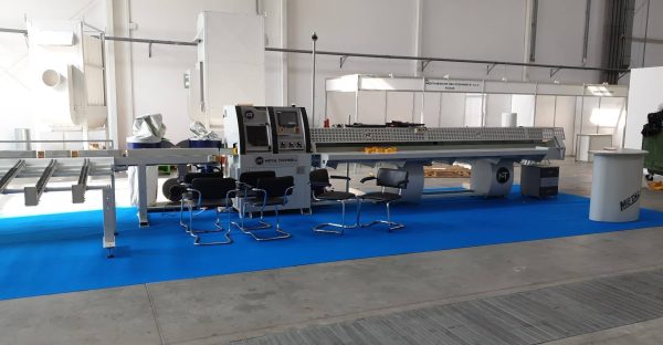 Stanowisko wystawiennicze z przemysłową maszyną Metal Technika na niebieskiej podłodze, otoczone krzesłami w hali wystawowej.