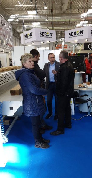 Cztery osoby na targach w hali przemysłowej przy stoisku firmy SERON z maszynami CNC, niebieska podłoga, białe bannery.