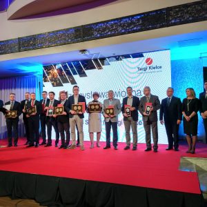 Grupa osób w garniturach stojąca na czerwonym dywanie na scenie podczas gali Targów Kielce, trzymająca złote nagrody, w tle e