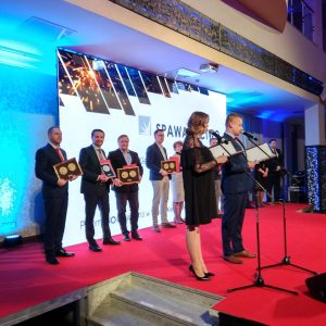 Ceremonia wręczenia nagród na czerwonym dywanie. Osoby w garniturach i sukniach trzymają złote puchary, dwie osoby stoją przy