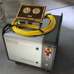 Urządzenie nLight Corona CFX-5000 z żółtymi przewodami i złotą skrzynką zawierającą dwie metalowe monety na brązowym tle.