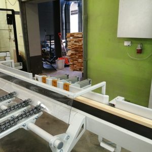 Hala produkcyjna z taśmociągiem. Na pierwszym planie urządzenie transportujące z rolkami, w tle zielona ściana, drewniane mat