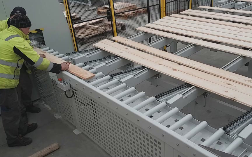 Pracownik w żółtej kamizelce bezpieczeństwa montuje drewniane deski na metalową konstrukcję z mechanizmem transportującym w h