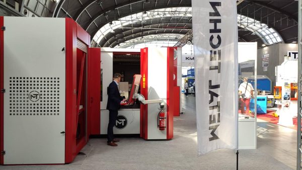Mężczyzna przy stanowisku wystawienniczym z czerwono-białymi panelami i logo MT na terenie hali wystawowej z metalicznym dach