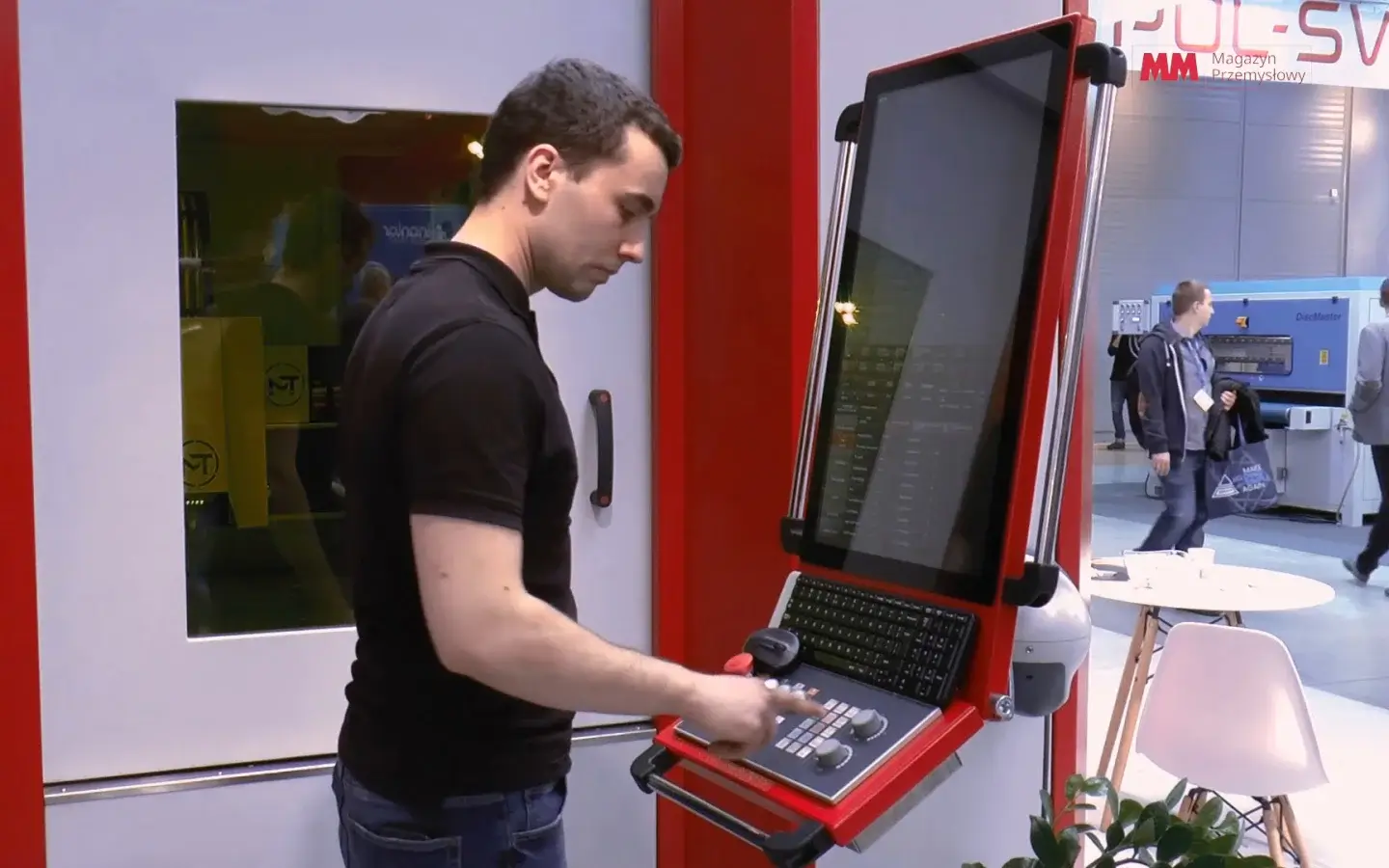 Mężczyzna w czarnej koszulce obsługuje czerwoną maszynę przemysłową z dużym ekranem dotykowym i klawiaturą na targu handlowym